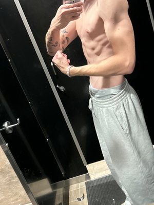 Shirtless man with tattoos flexing in mirror, wearing gray sweatpants, indoor setting.