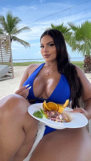 Woman in blue bikini top holding plate of food on beach, palm trees in background.