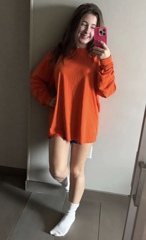 Woman in orange oversized shirt and white socks, taking mirror selfie indoors.