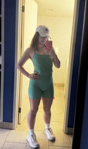 Woman in green crop top and shorts, white cap, sneakers, taking mirror selfie in bathroom.