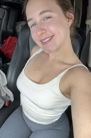 Woman in white tank top and gray leggings sitting in car, smiling at camera.
