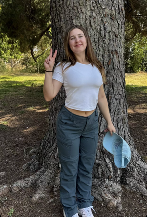Woman in white crop top and gray pants leaning against tree in park, holding cap.