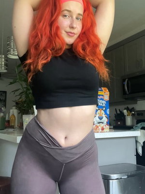 Woman with bright red hair in black crop top and gray leggings posing in kitchen.