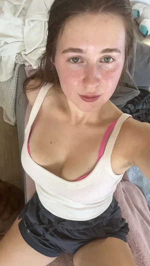 Woman in white tank top and black shorts sitting on bed, looking at camera.