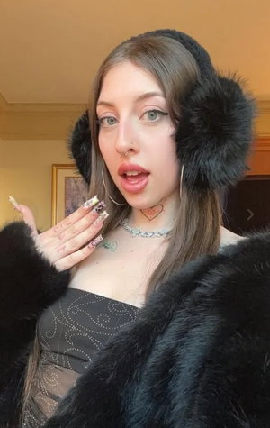 Woman with brown hair wearing black fur coat and earmuffs indoors.