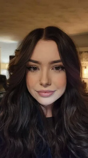 Woman with long dark hair and makeup, wearing dark top, indoors.