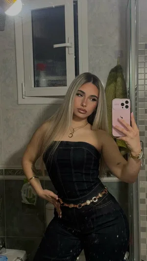 Blonde woman taking mirror selfie in bathroom wearing black top and jeans.
