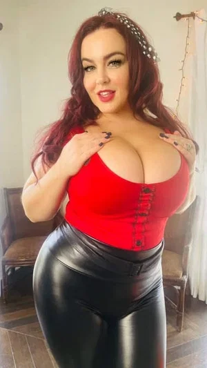 Woman with red hair in red corset top and black leather pants posing indoors.