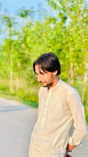 Man in light-colored traditional shirt standing outdoors near green trees.