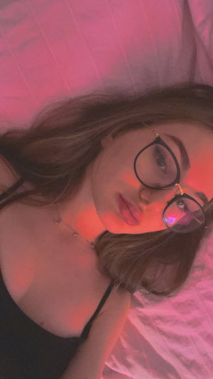 Woman with glasses lying on bed, wearing black top, pink lighting.