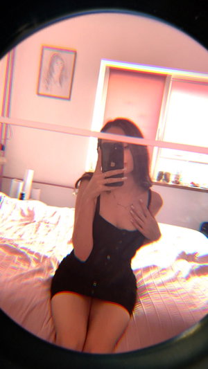 Woman in black dress sits on bed, taking mirror selfie in bedroom.