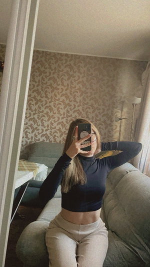 Woman in black crop top and beige pants sits on couch, taking mirror selfie in bedroom.
