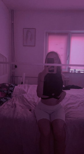Woman in black crop top and light shorts sits on bed in dimly lit room with pink hue.