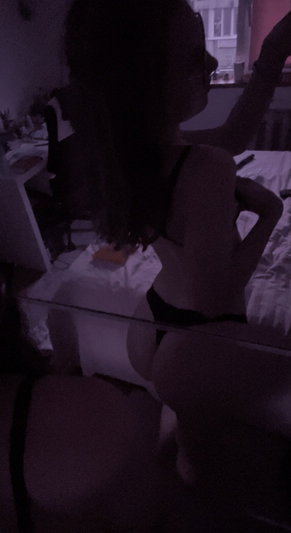 Woman in black lingerie sits on bed, back to camera, in dimly lit room.