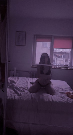 Woman in black lingerie kneeling on bed, taking mirror selfie in dimly lit bedroom.