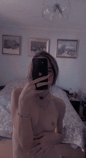 Woman with bare chest and covered breasts takes mirror selfie in bedroom.