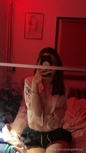 Woman in white shirt and black shorts sitting on bed, taking mirror selfie in red-lit room.