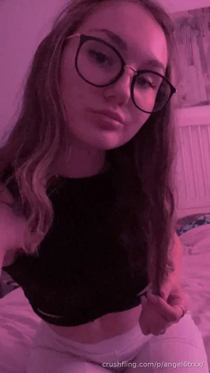 Woman with long hair and glasses in black crop top and white pants, sitting on bed in purple-lit
