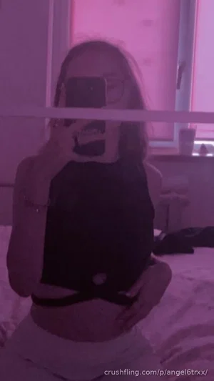 Woman in black crop top and light pants taking mirror selfie in bedroom with pink lighting.