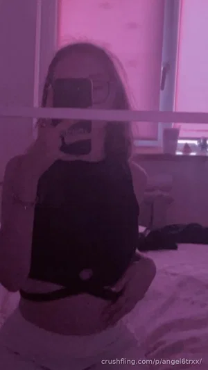 Woman in black crop top and light pants taking mirror selfie in bedroom with pink lighting.