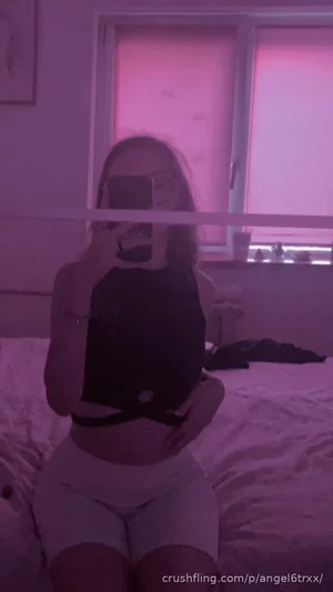 Woman in black crop top and light shorts sits on bed, taking mirror selfie in pink-lit room.