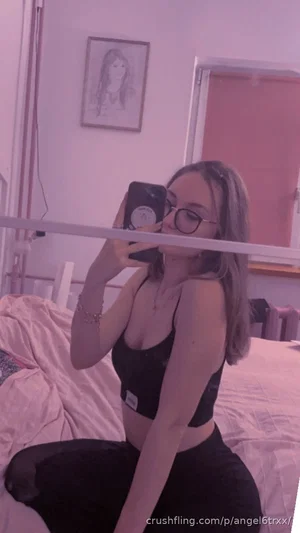 Woman in black crop top and pants sitting on bed, taking mirror selfie in bedroom.