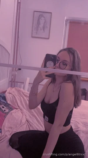 Woman in black crop top and pants sits on bed, taking mirror selfie in bedroom.