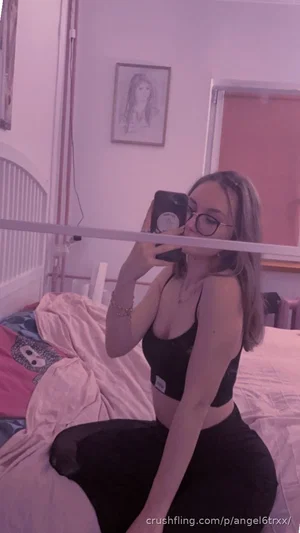 Woman with glasses in black bra and pants sits on bed, taking mirror selfie in bedroom.