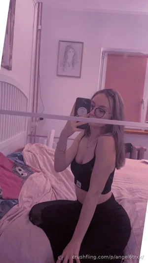 Woman with glasses in black crop top and pants sitting on bed in bedroom, taking mirror selfie.