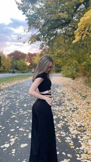 Woman in black crop top and long pants standing on leaf-covered path outdoors.