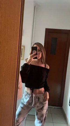 Woman in off-shoulder top and tie-dye pants takes mirror selfie indoors.