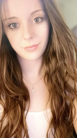 Woman with long brown hair wearing white top, indoors, close-up selfie.