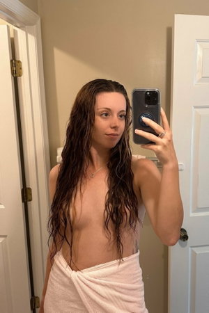 Woman with wet hair wrapped in towel, taking mirror selfie in bathroom.