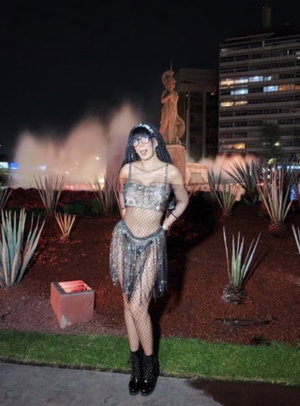 Woman in sparkly outfit and fishnet stockings posing outdoors at night near a fountain.