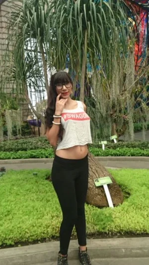 Woman with long dark hair in a crop top and black leggings posing outdoors near plants.