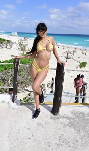 Woman in yellow bikini posing on beachside walkway, wearing glasses and black sneakers.