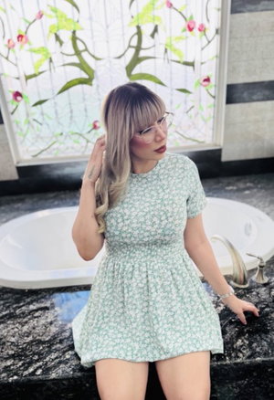 Woman with blonde hair and glasses in a floral dress sitting on a bathroom counter.