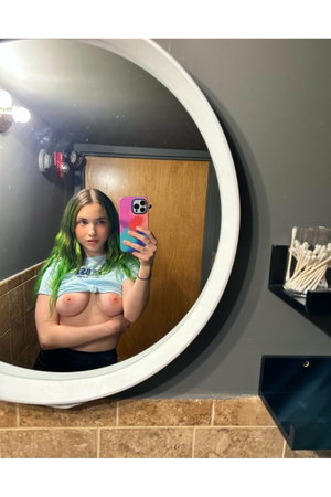 Green-haired woman taking mirror selfie in bathroom wearing tied shirt.