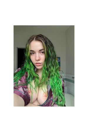 Woman with green wavy hair in open purple satin top, indoor.