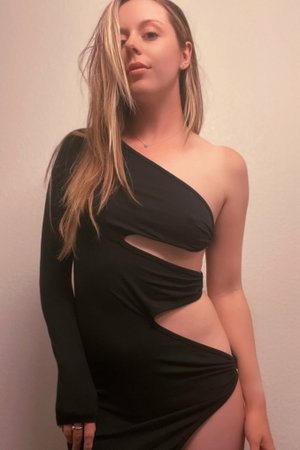 Woman in black one-shoulder dress with cutouts, standing against plain wall.