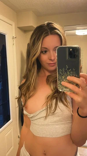 Woman with long blonde hair in a light crop top taking mirror selfie in bathroom.