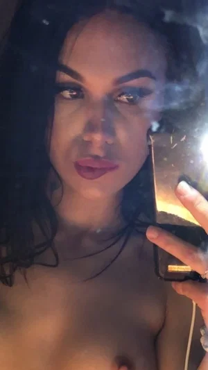 Woman with dark hair and makeup taking mirror selfie, bare chest visible, holding phone.