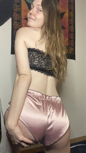 Woman in black lace top and pink satin shorts posing indoors, back to camera.