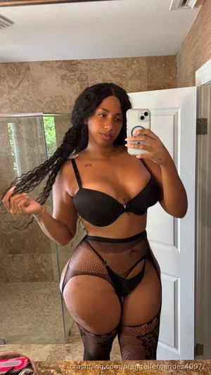 Woman with long dark hair in black lingerie and fishnet thigh-highs taking mirror selfie in