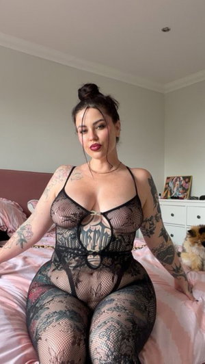 Tattooed woman in black fishnet bodysuit on bed.