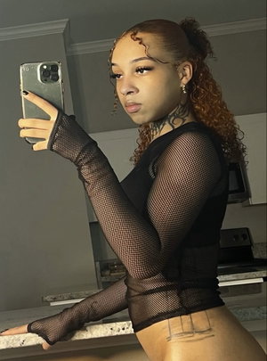 Woman with curly hair in black mesh top taking mirror selfie in kitchen.