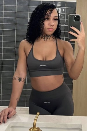Woman with curly hair in gray sports bra and leggings takes mirror selfie in bathroom.
