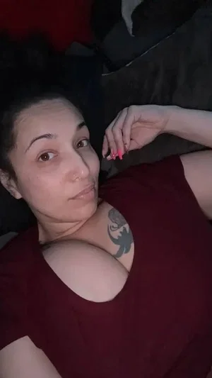 Woman with tattoo on chest wearing red top, lying down in dim lighting.