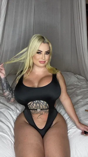 Blonde woman in black bodysuit on bed with tattoos.