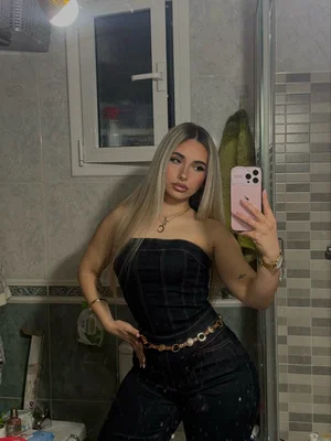 Blonde woman taking mirror selfie in bathroom wearing black top and jeans